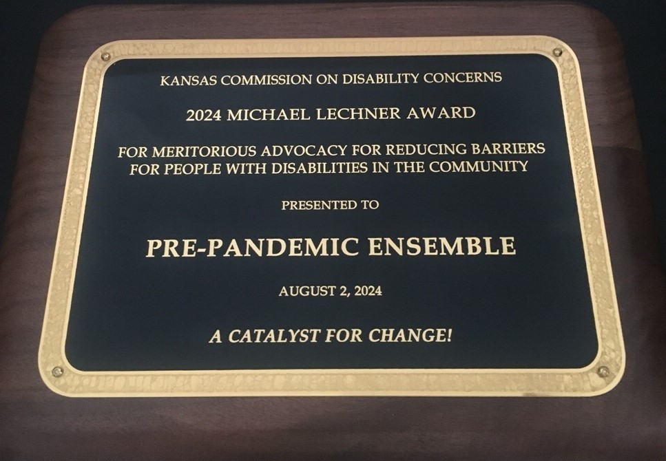 Photograph: Wooden plaque with black panel and gold border and writing. Text: Kansas Commission on Disability Concerns. 2024 Michael Lechner Award for Meritorious Advocacy for Reducing Barriers for People with Disabilities in the Community. Presented to Pre-Pandemic Ensemble. August 2, 2024. A Catalyst for Change!