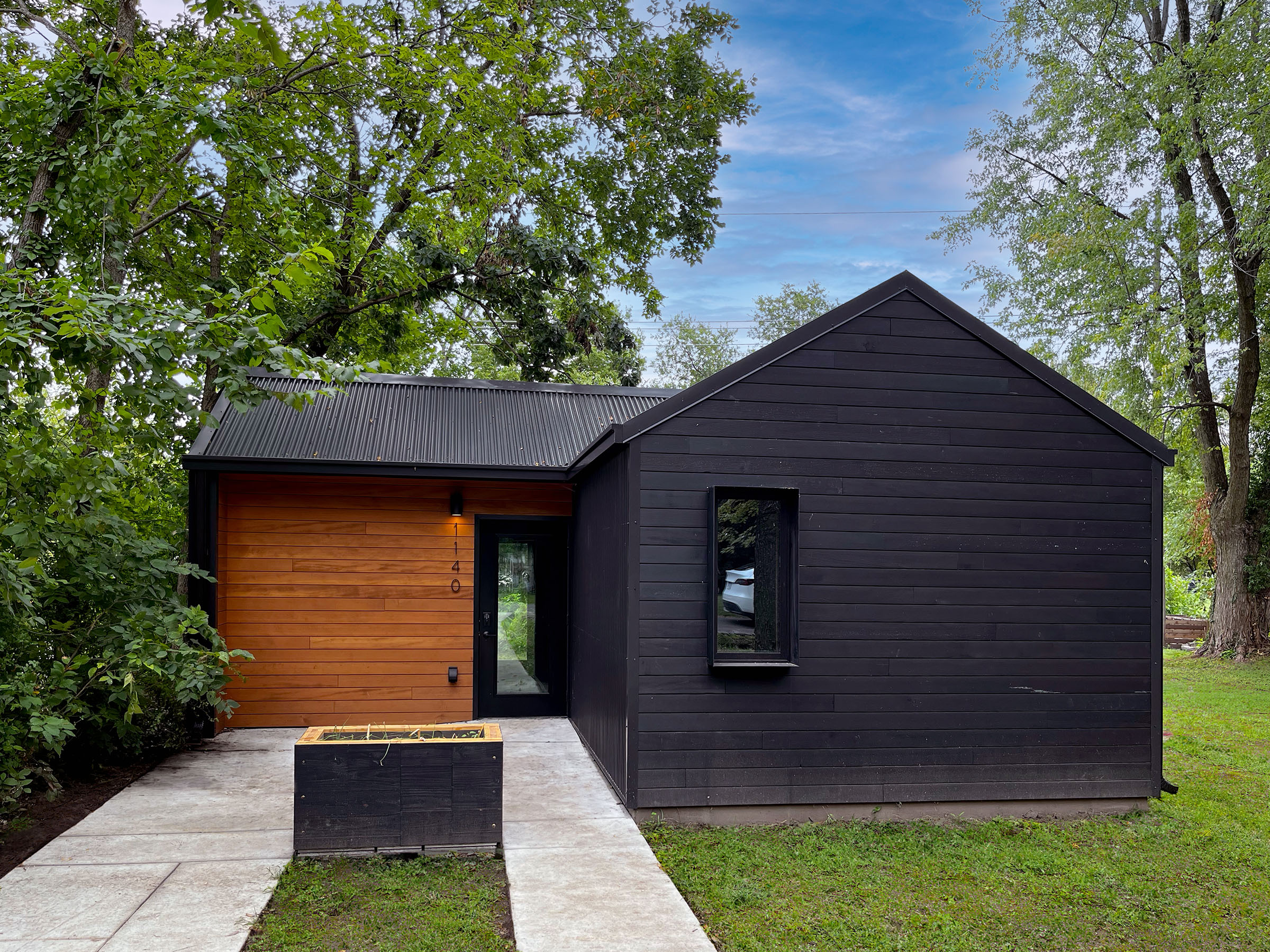 Exterior view of Phoenix House, a small modern house with clad in black and contrasting natural wood siding, surrounded by lush greenery.