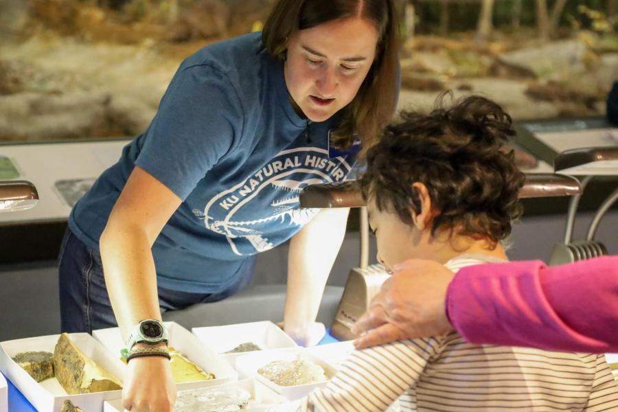 Kelly Pfeiler points to a plant fossil specimen on a table while explaining it to a young museum visitor.