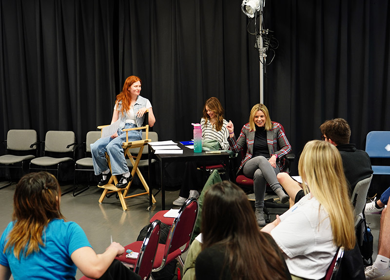 Guest artist seated in a studio room where a student sits in a director's chair. Others are seated around her.