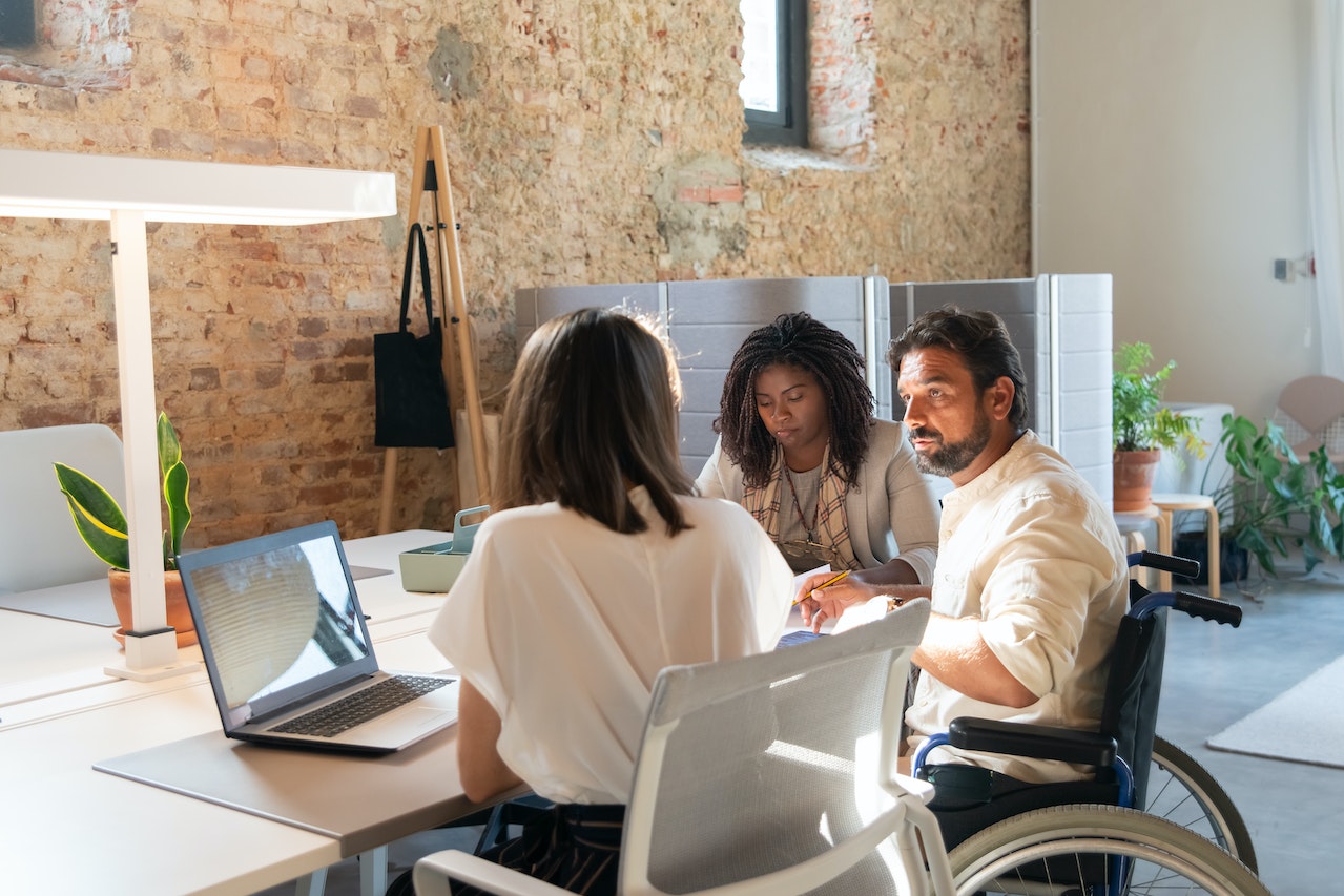 "Two woman sit at a table with an open laptop as they interact with a man who is seated in a wheelchair"