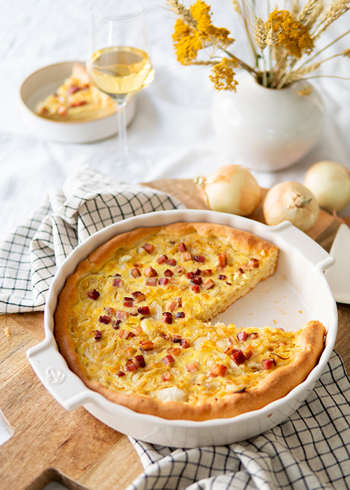 A freshly baked onion pie with pieces of bacon on top, served on a wooden cutting board beside a glass of white wine and garnished with yellow flowers in a vase.