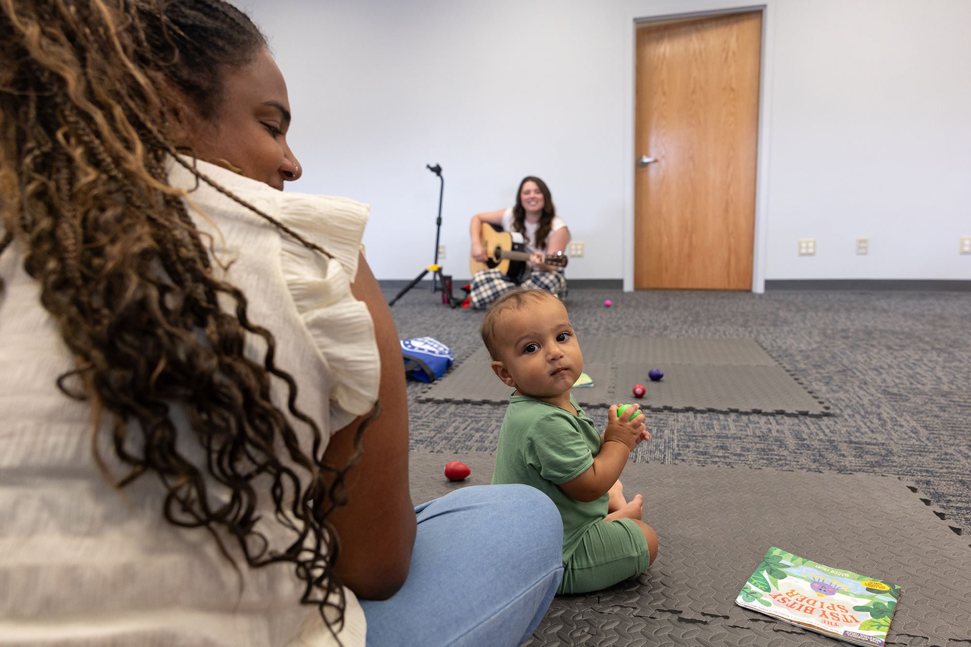 A baby sitting in front of their mother looks back while listening to a musician playing at the front of the room