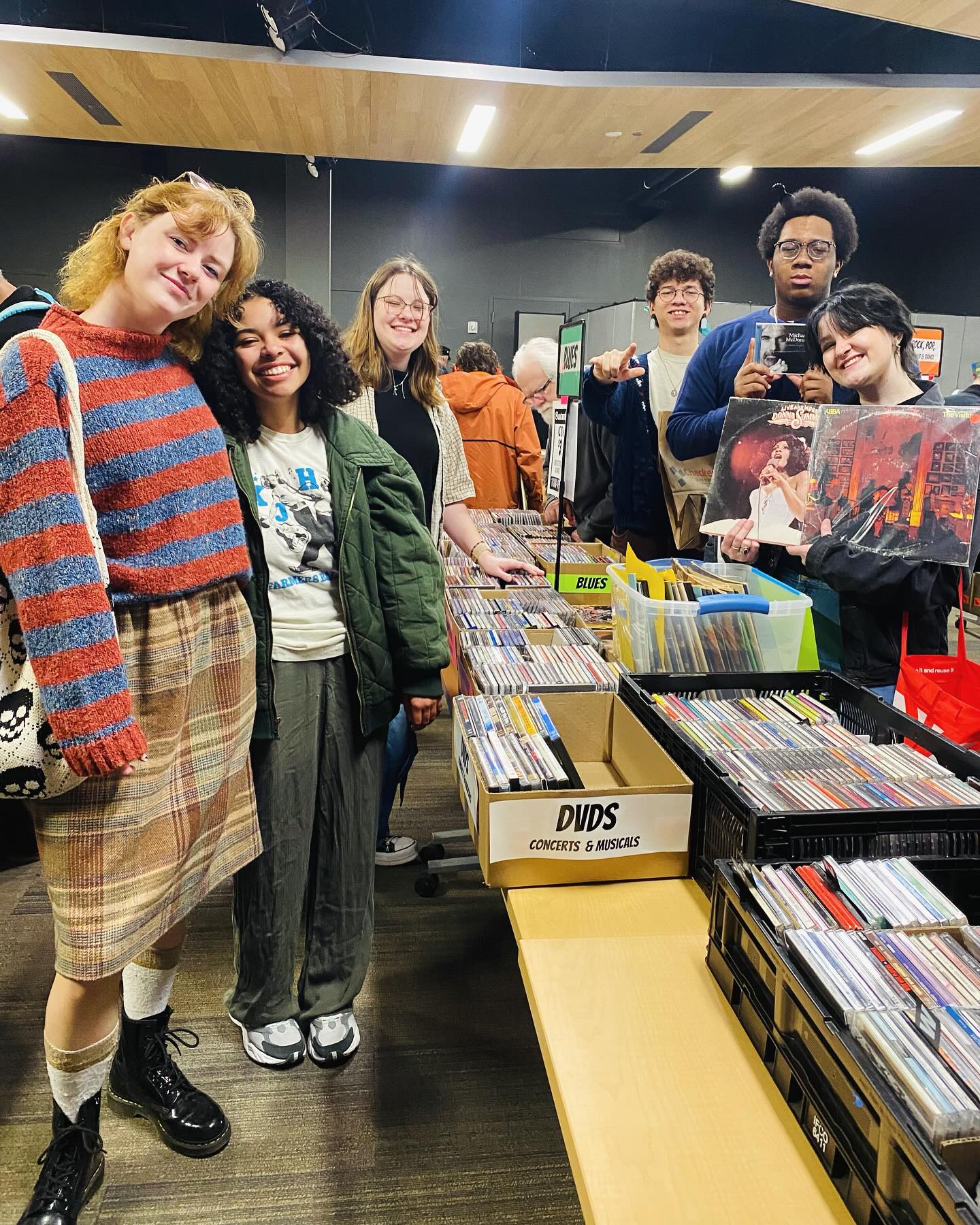 Six volunteer students from KJHK stand in front of several boxes of CDs and DVDs for sale during Audio-Reader's February audio equipment sale at the Lawrence Public Library