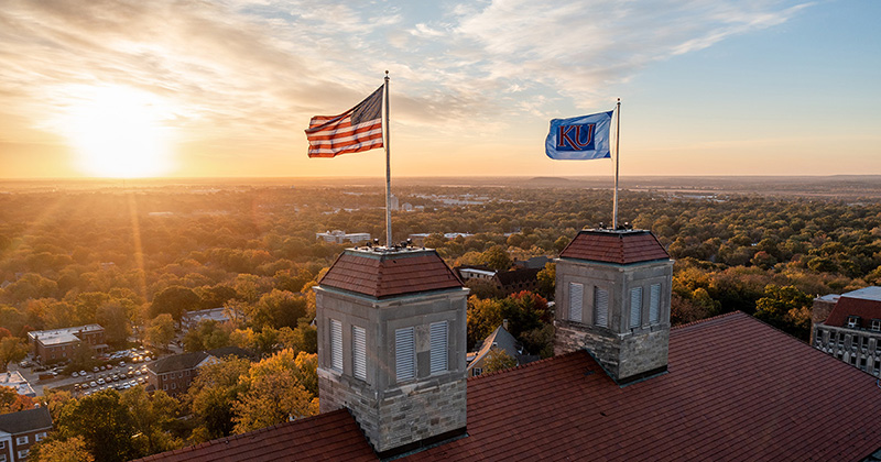 The American flag and KU flag atop Fraser Hall wave in the wind as the sun rises in the background.