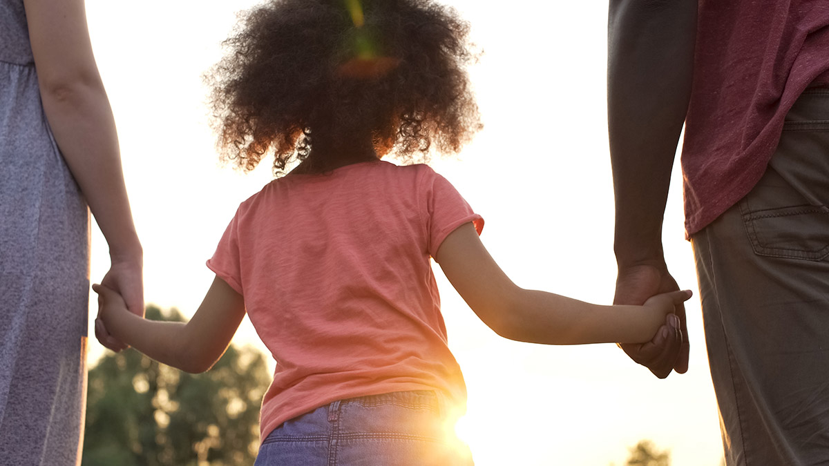 An image of parents holding hands with a child as the sun sets in the background of the happy family.