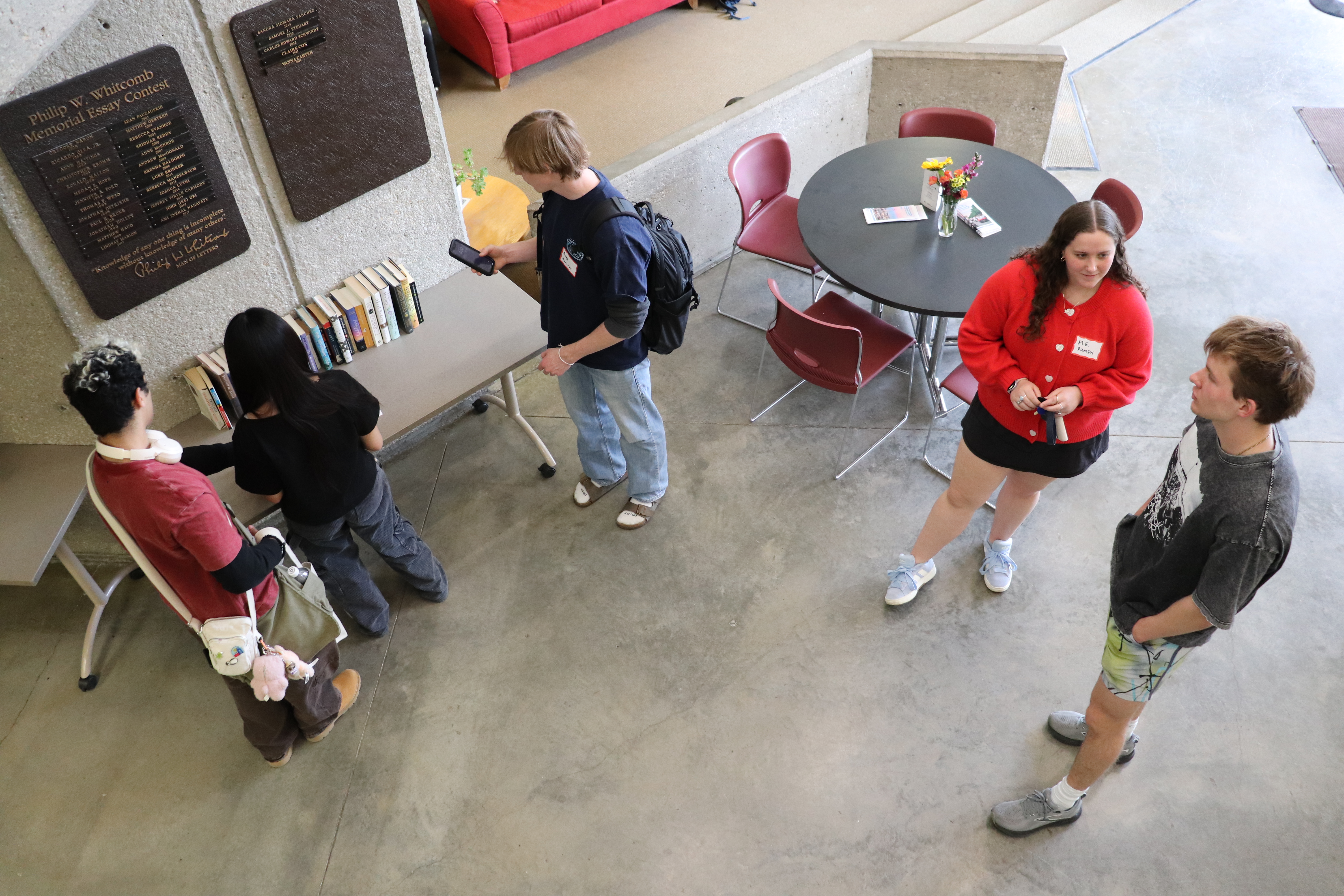 Three students look at books on table; two stand in conversation