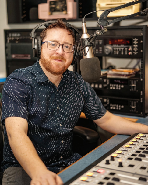 Nick Carswell sits in front of a recording microphone in a recording studio with a soundboard at his fingertips. Nick wears glasses, has reddish-brown short hair and a matching full beard.