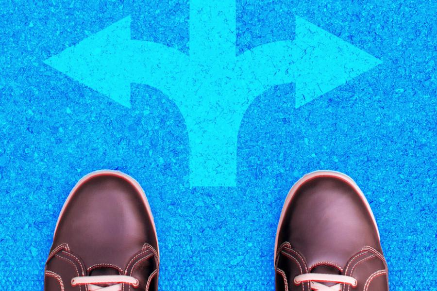 A pair of feet stand on pavement at crossroads marked by painted white arrows pointing in different directions