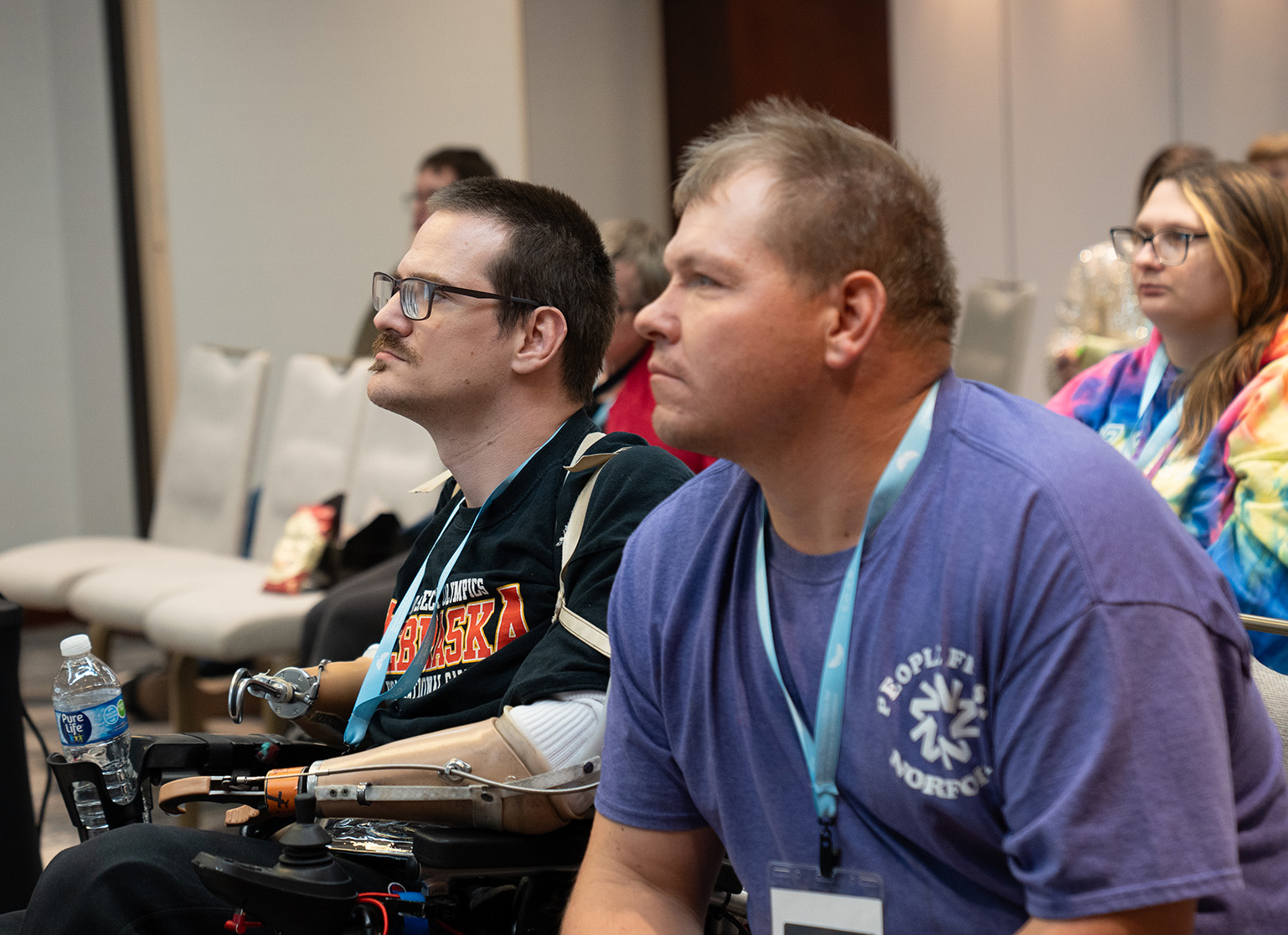 "A man with blond hair wearing a purple shirt leans forward in his chair while sitting next to a man with glasses and a black shirt and prosthetic arms while seated in a wheelchair as they both listen as audience members"