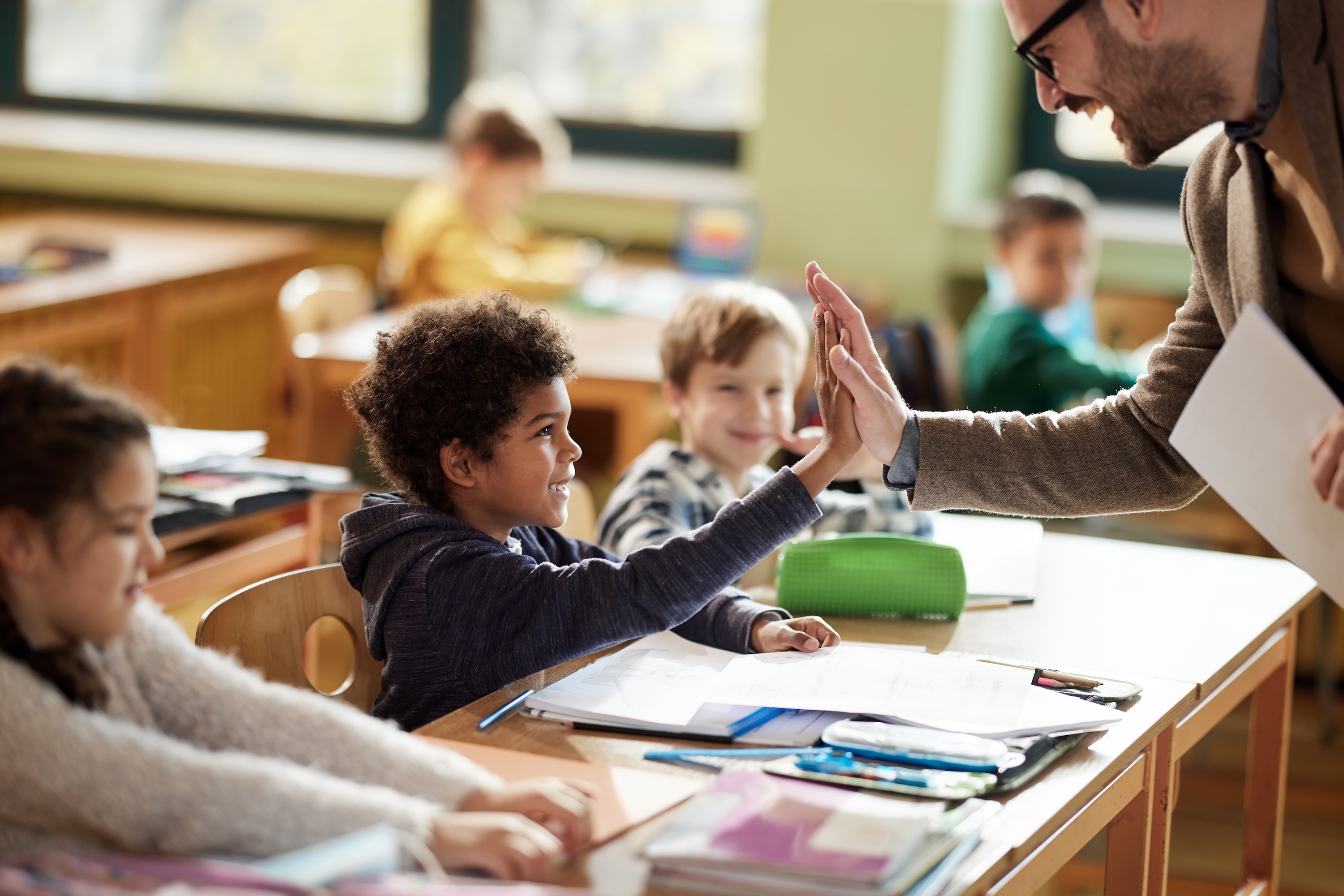 teacher gives student sitting at a desk with other students a high five and smiles