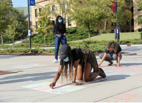 A masked woman kneels, drawing with chalk on a sidewalk