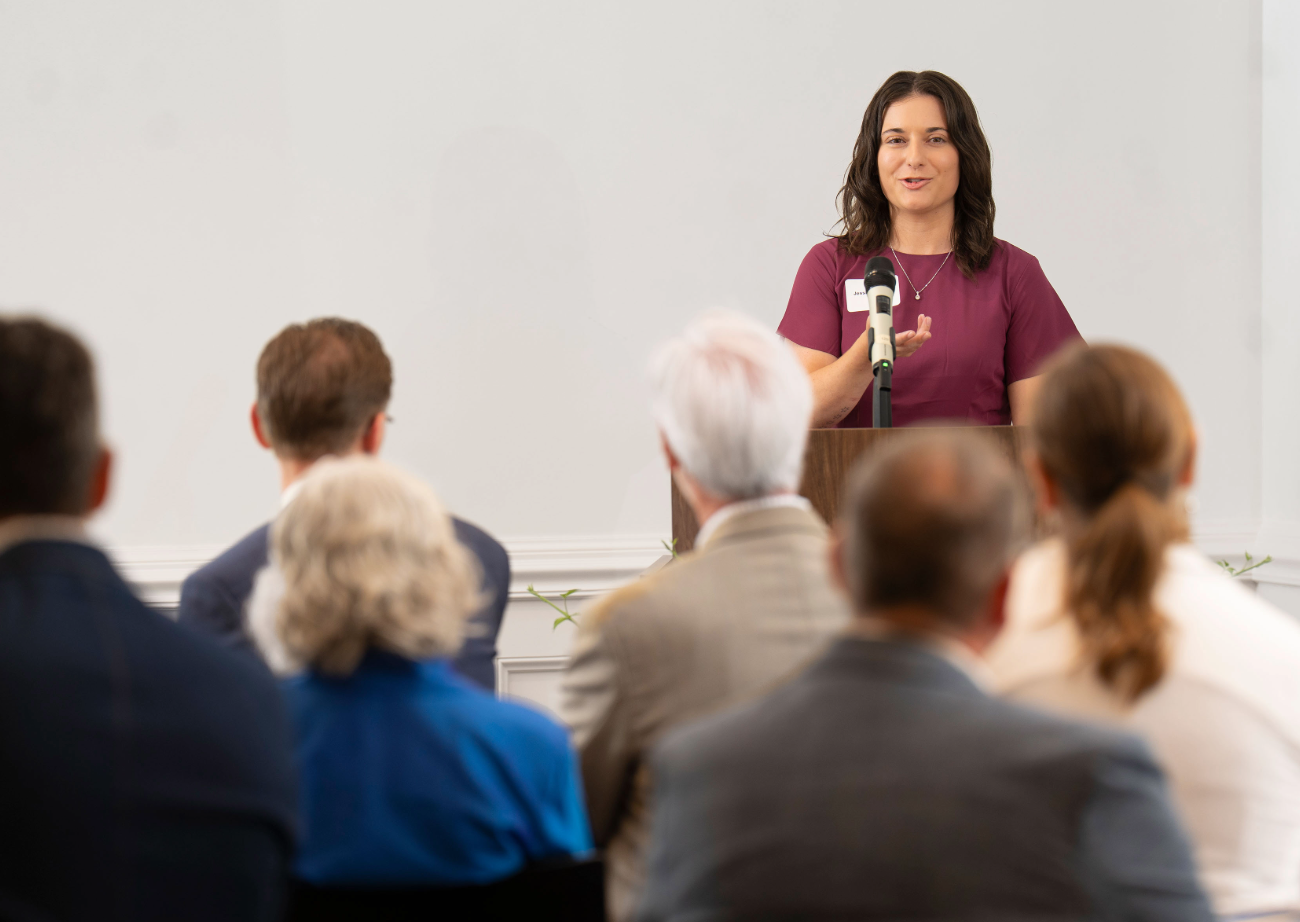 Jessica Powers speaks to a crowd while standing at a podium at the head of a conference room
