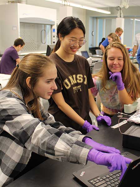 Three students in a laboratory setting, wearing protective gloves and engaging with their experiment. They are focused on the task at hand with expressions of curiosity and concentration.