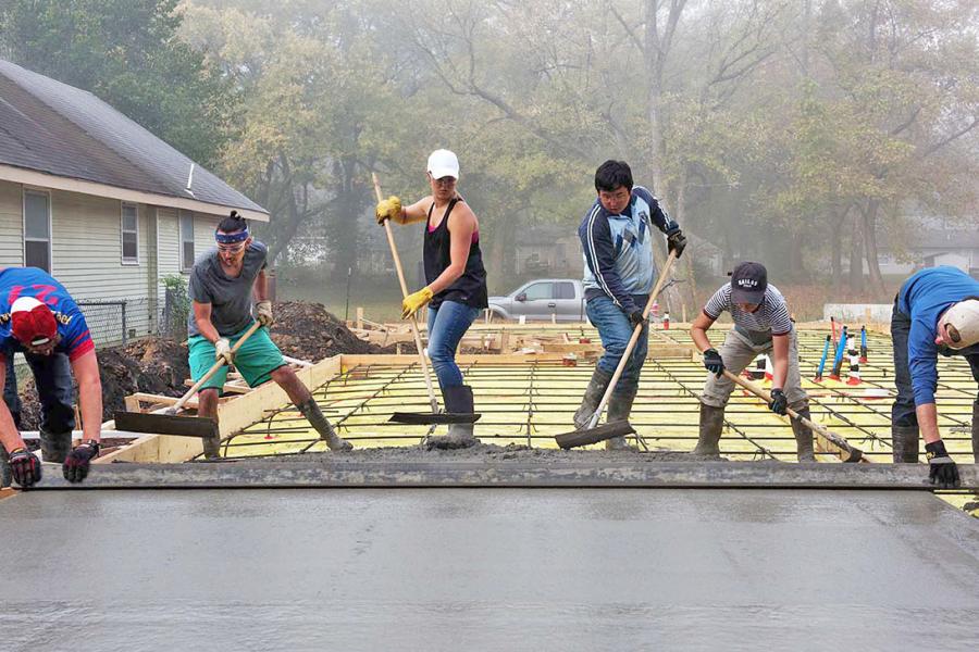 Group of students in a line, cooperatively using hand tools to smooth out a pad of concrete.