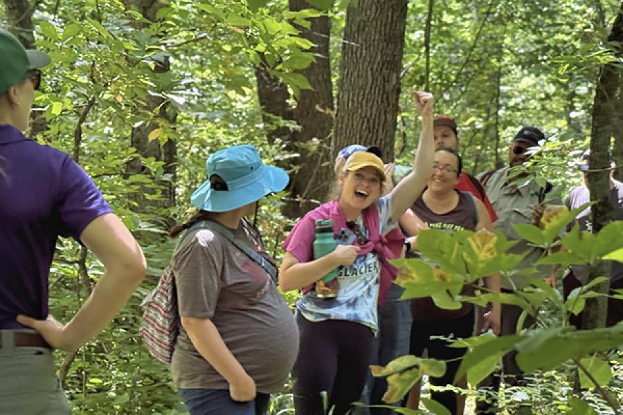 Group of eight people travel along forest path. One person raises hand and shouts while others smile. The group leader, in the uniform of a forester, with polo shirt, hiking pants and cap, has turned to look toward the group.