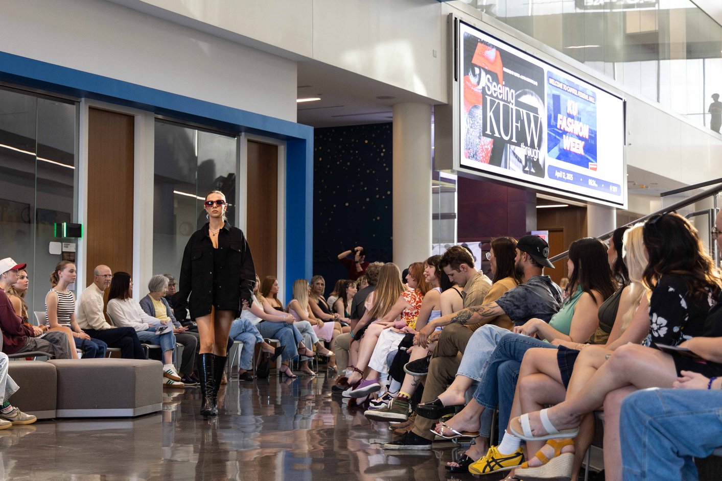 A female model wearing a black jacket and dress with black boots walks the runway during a fashion show.