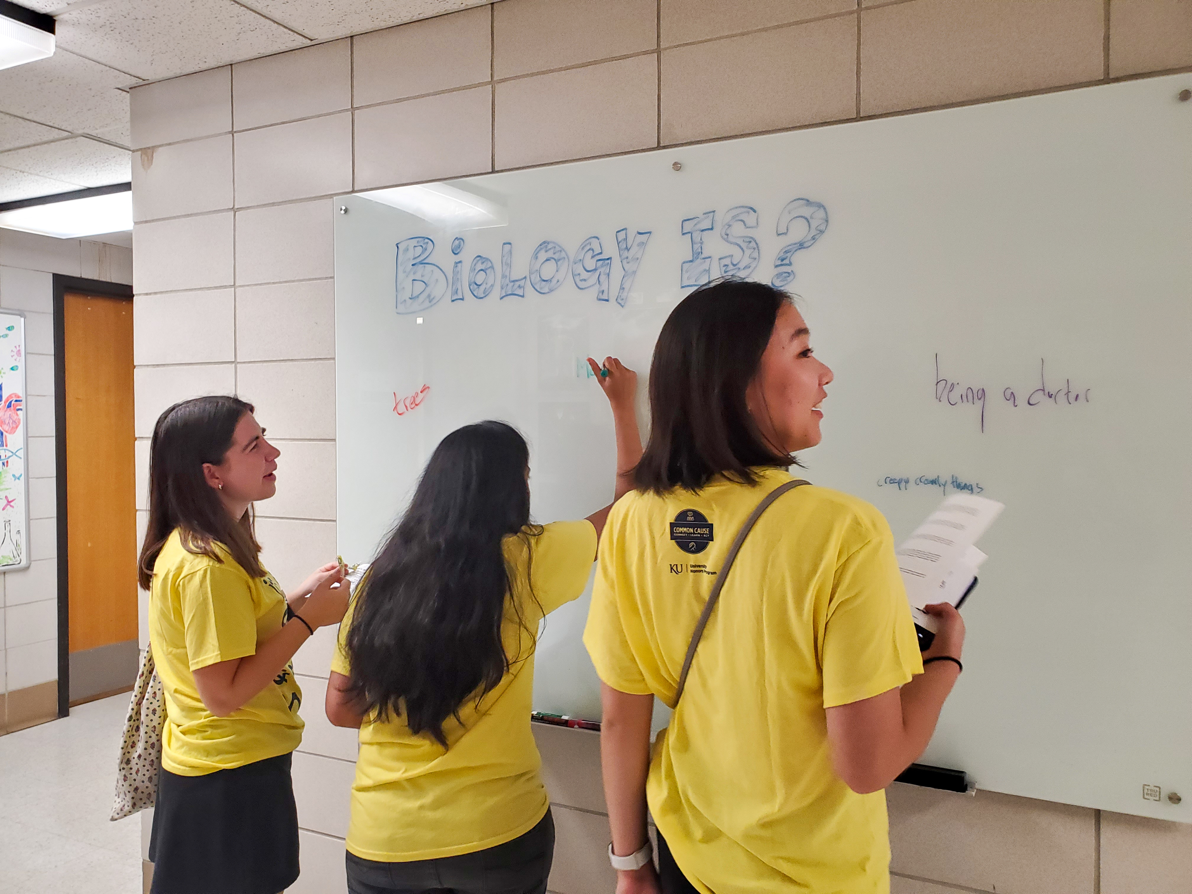 three students in matching t shirts answer the question "Biology is" on a white board