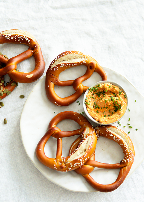 Three soft pretzels served with a side of mustard dip on a white marble plate, garnished with salt and herbs, on a light linen backdrop.