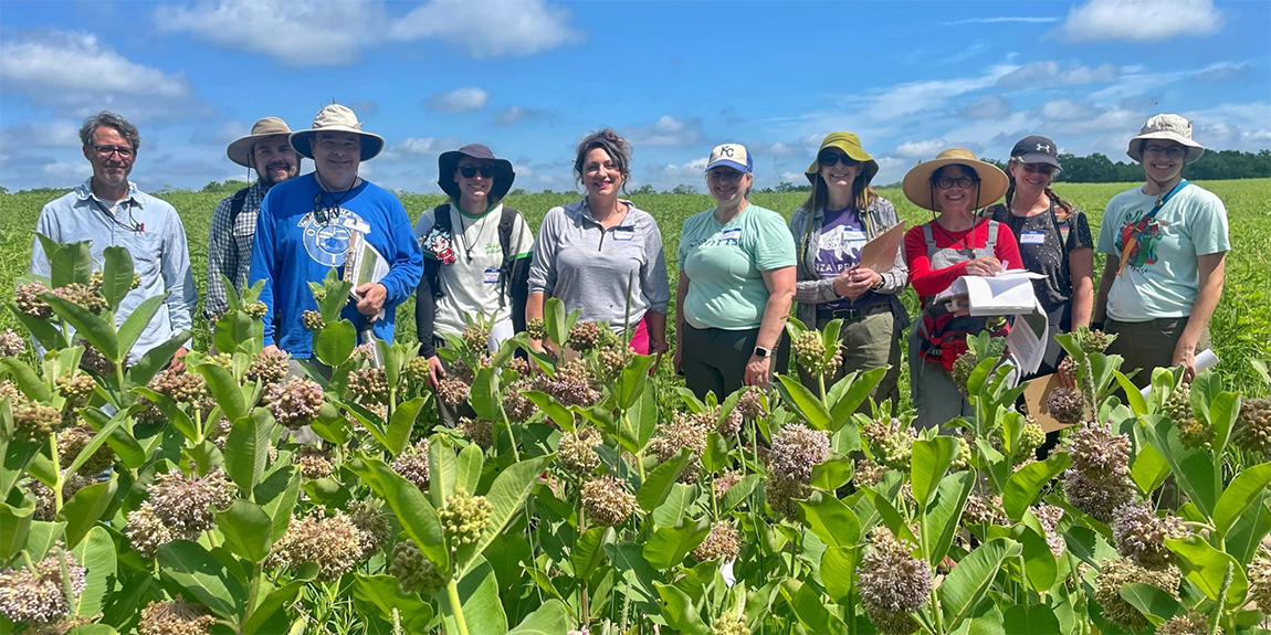 Group of people standing in a line facing camera in prairie setting with milkweed blooming in foreground.