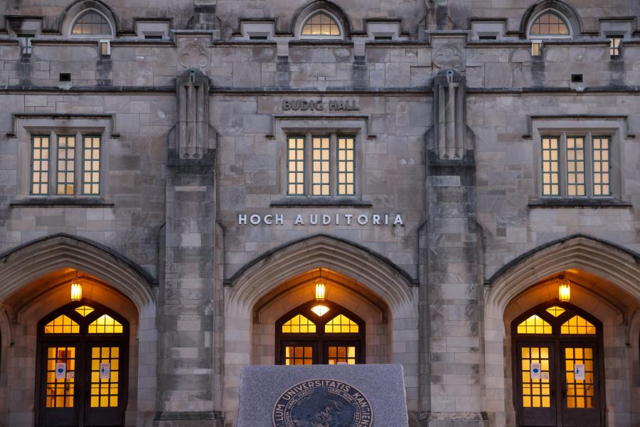 Exterior view of Budig Hall and Hoch Auditoria at dusk with illuminated windows and a visible monument in the foreground.