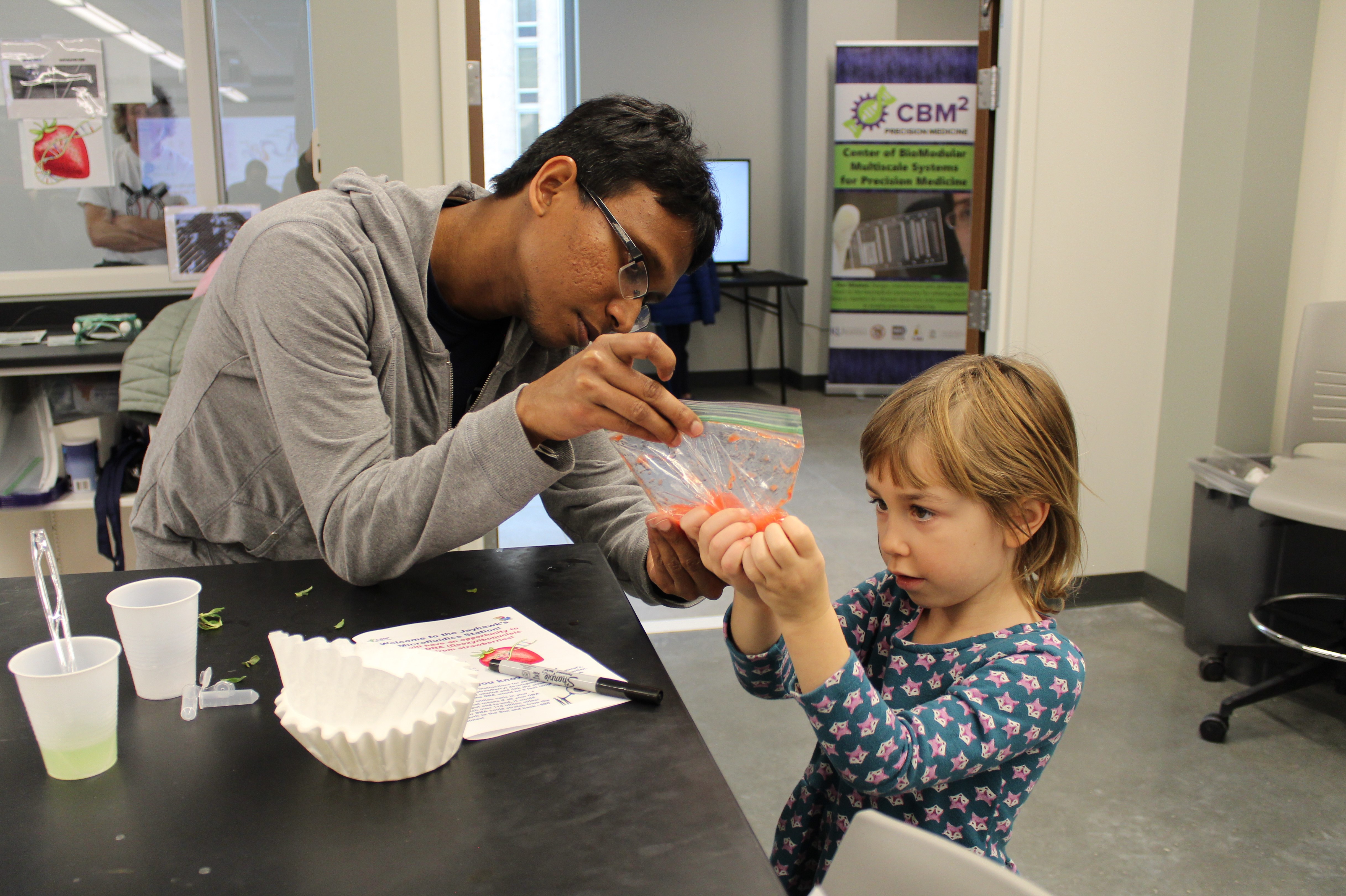 Helper assisting a child with a plastic bag containing a red substance.