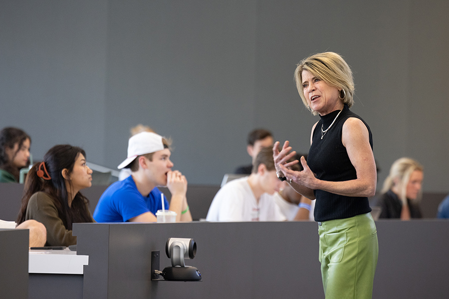 A woman wearing a black shirt and green pants teaches in front of a classroom.