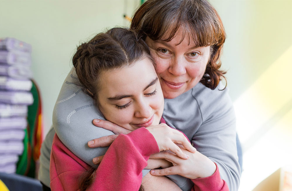 A woman with brown hair is looking at the camera as she hugs a child who has closed her eyes and is slightly smiling.