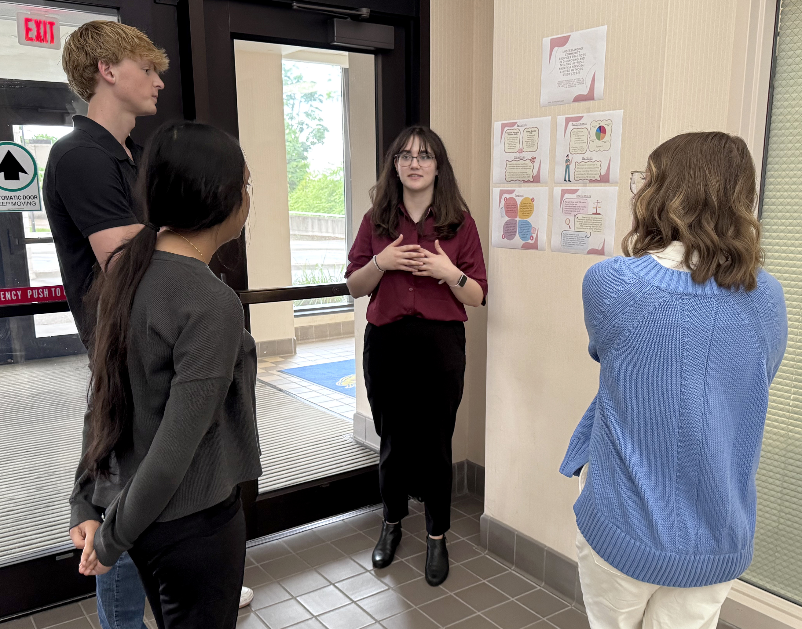 A student wearing a burgundy shirt and black pants speaks to three individuals while standing next to research papers taped to the wall.