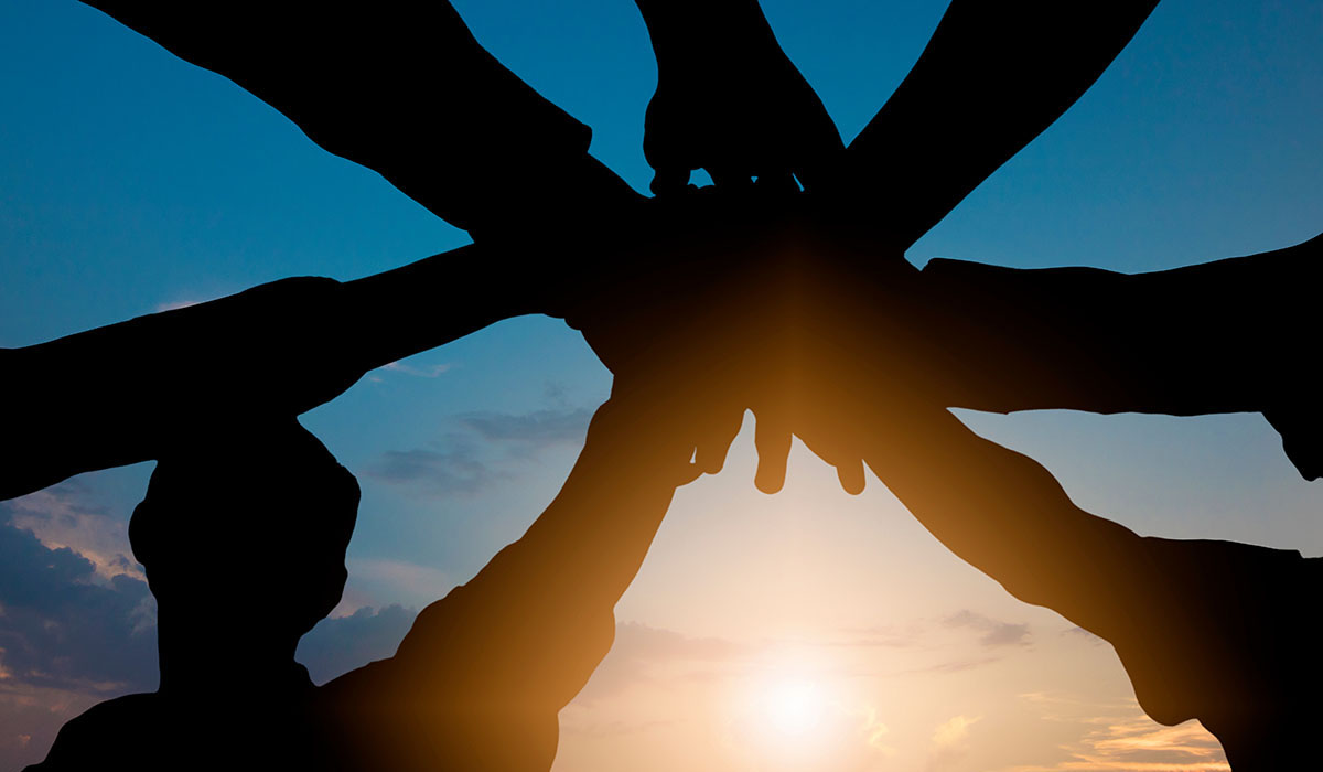 A group of hands are joined before the backdrop of a sunset.