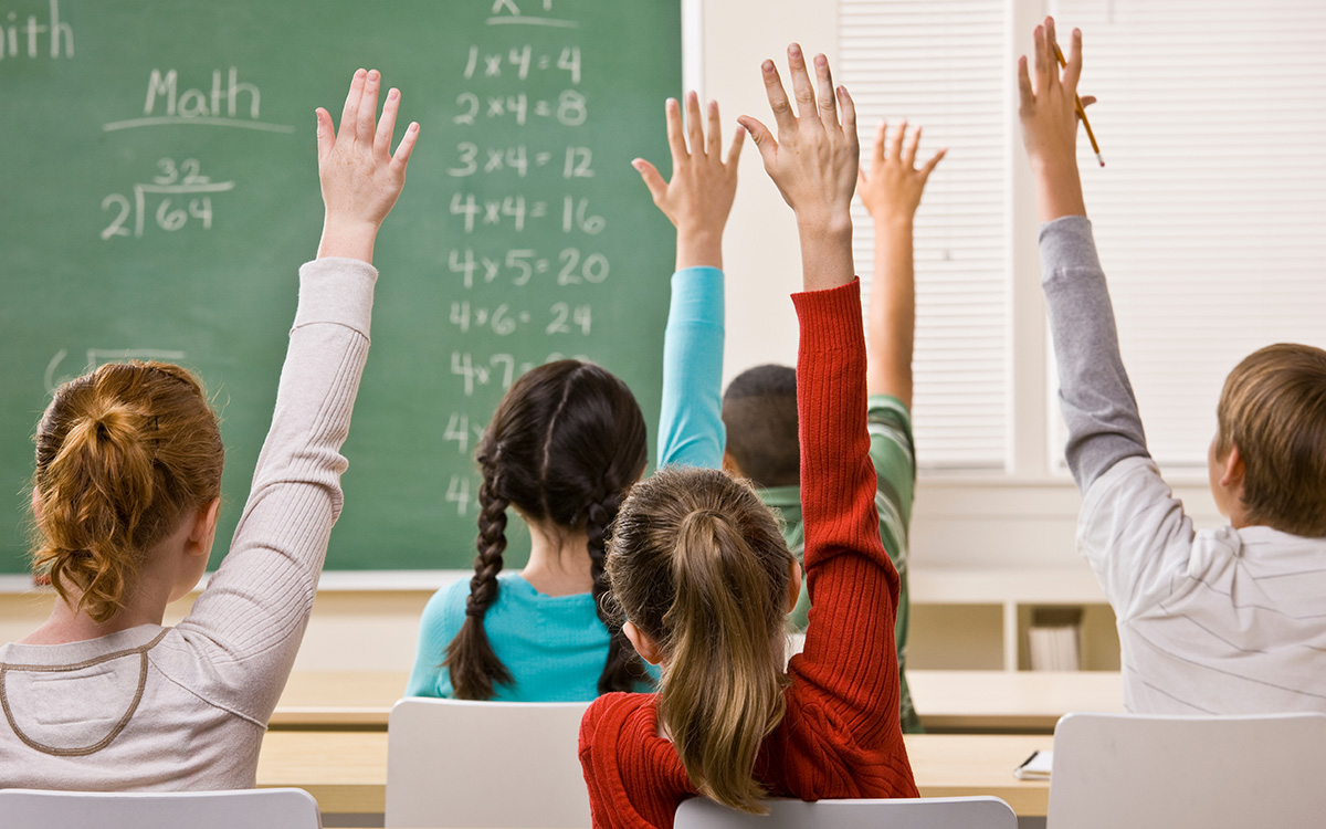 Students raise their hands in a classroom with math formulas written on chalkboard.