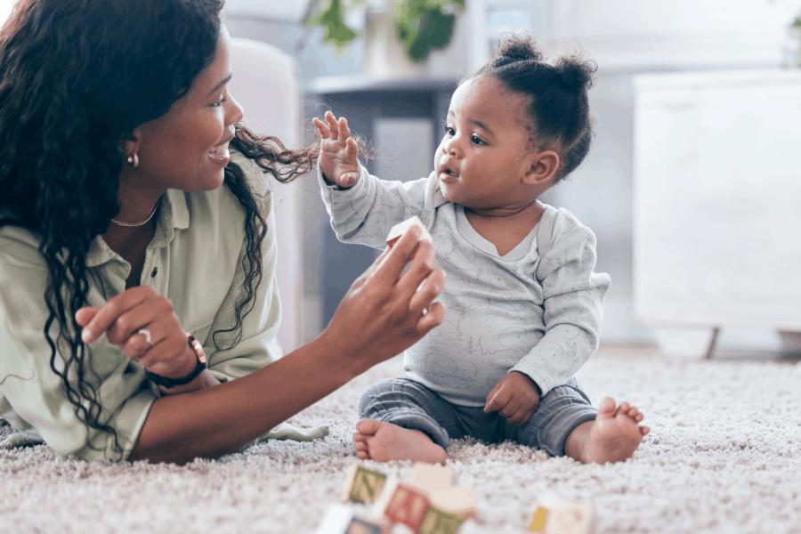 A baby sitting on a carpeted floor reaches toward her mother, who is smiling at her and holding a toy block.