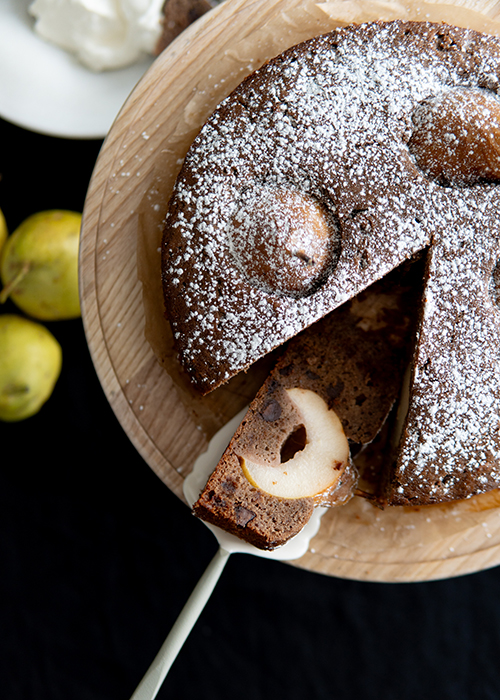 A top-down view of a sliced pear cake sprinkled with powdered sugar, served on a wooden plate with a side of whipped cream. Fresh pears accompany the plate.