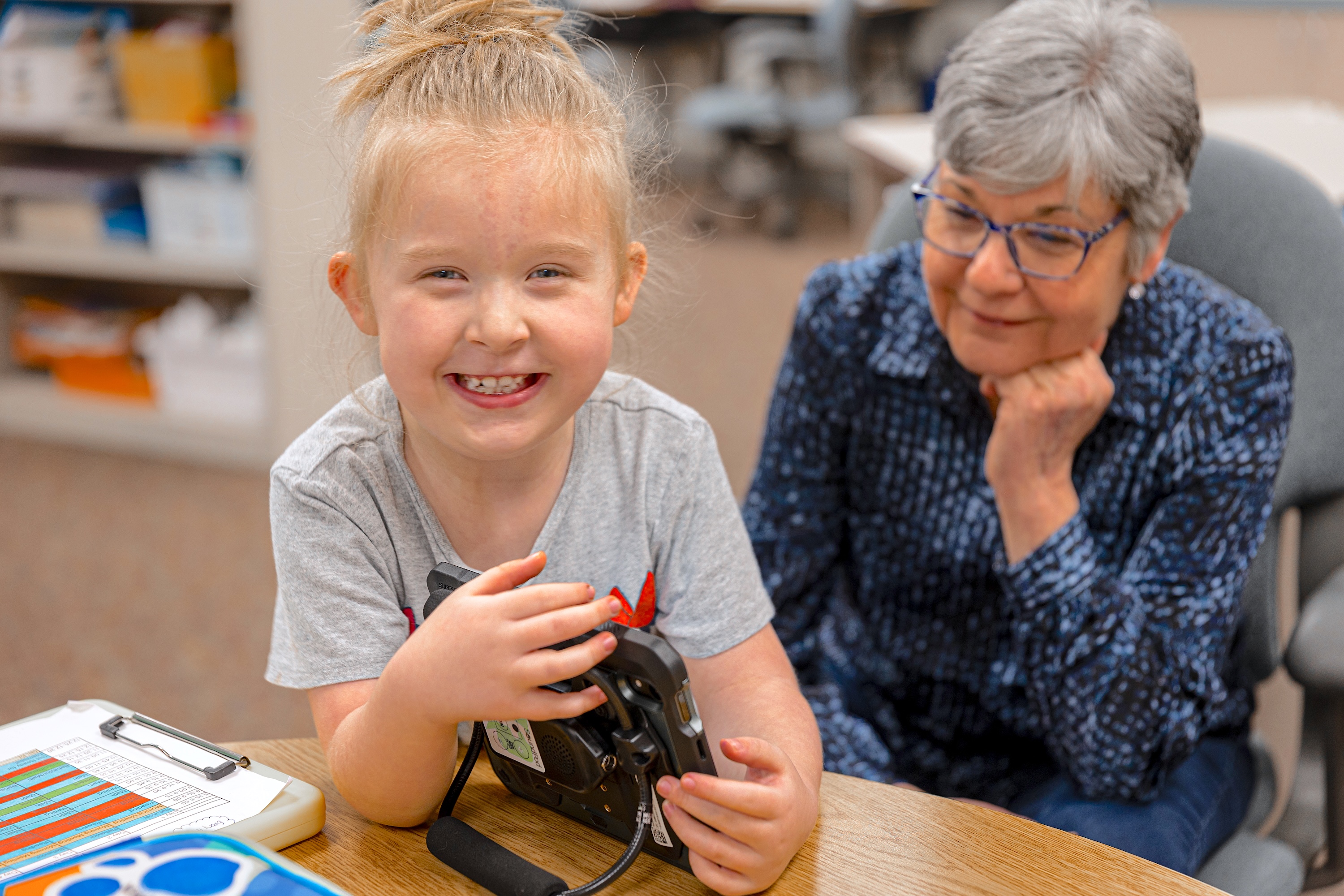 young student holding assistive communication device smiling at camera with educator sitting behind the student