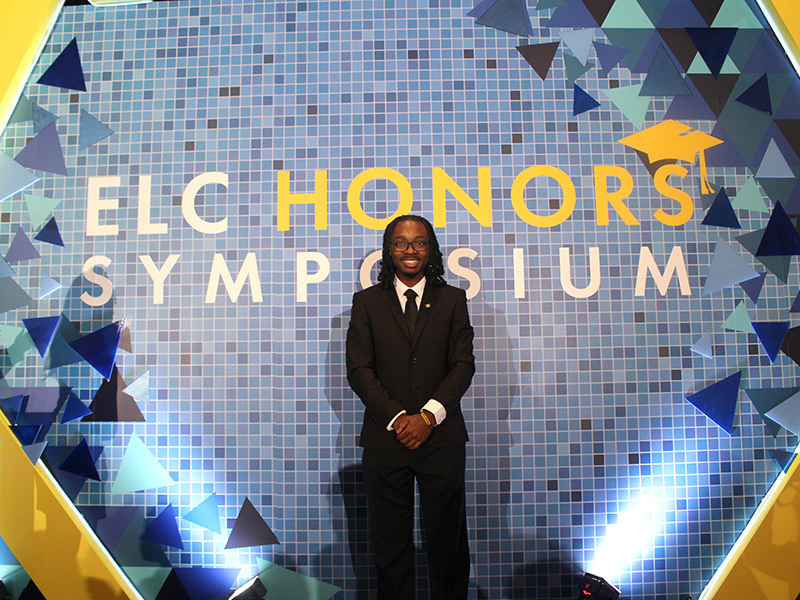 An individual in a suit stands in front of a geometric-patterned backdrop with the text "ELC Honors Symposium."