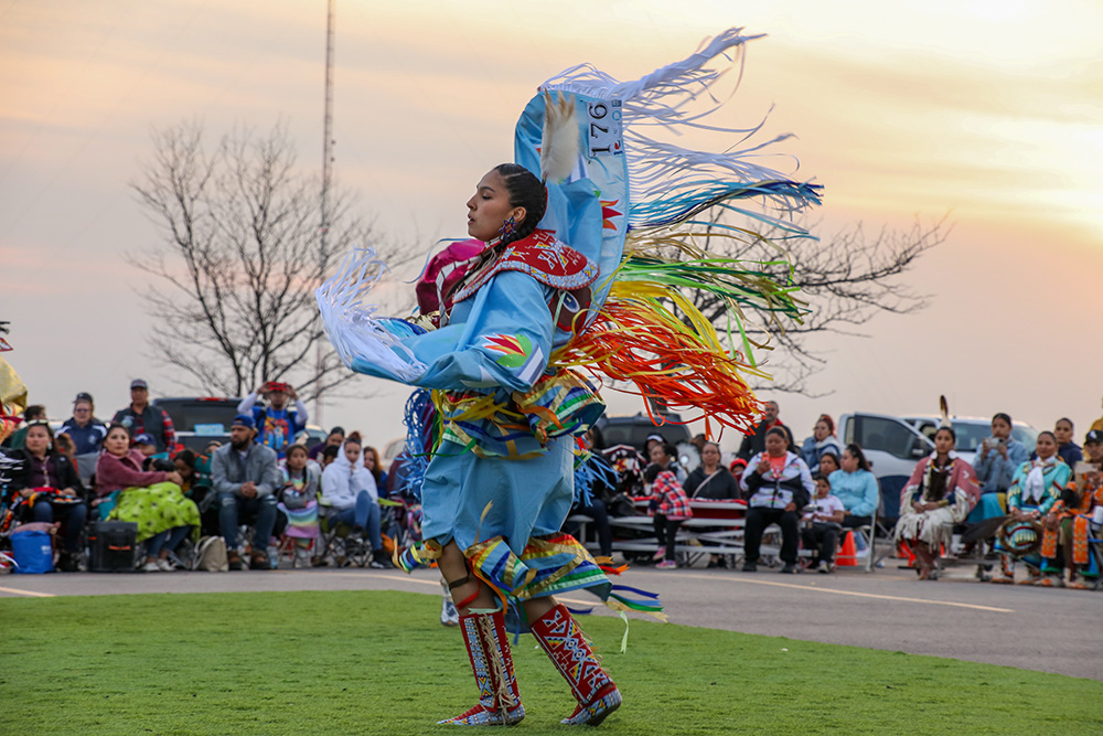 An individual with long, dark braids and bright, multicolored attire dances with seated crowd in background. Credit: Laura Kingston.