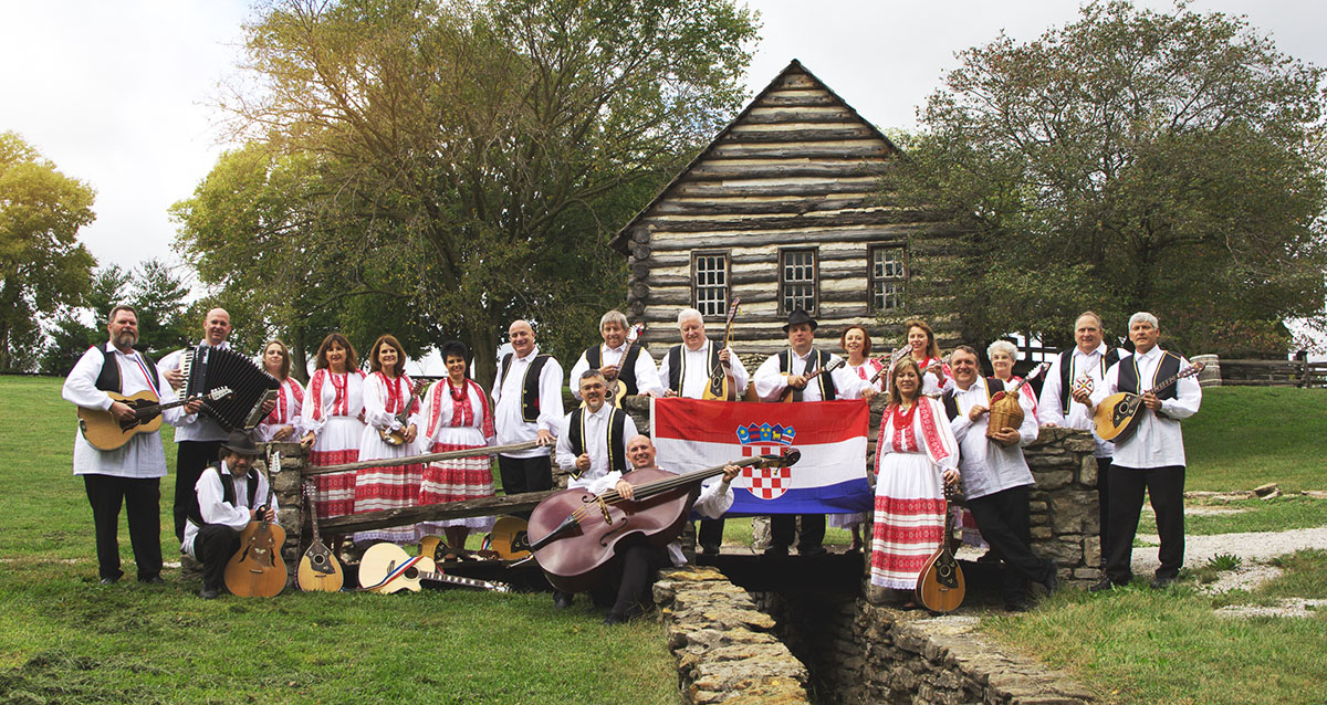 Outdoor group photo of Hrvatski Običaj, a 20-piece Croatian band, in front of a log cabin.