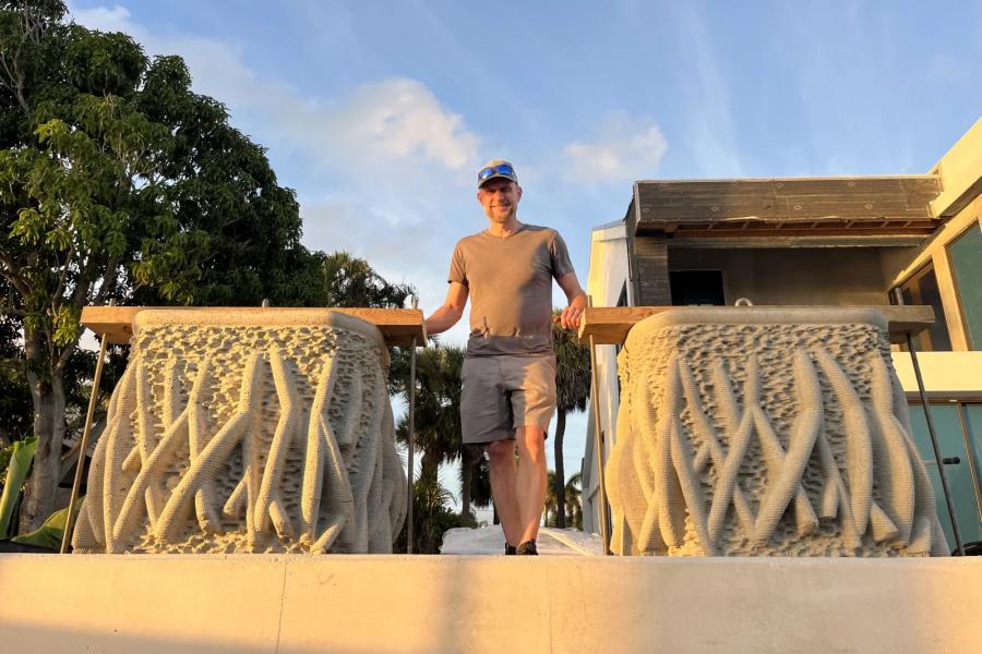 Keith Van de Riet stands on a waterfront walkway between two mangrove inspired concrete seawall planters, featuring textured surfaces that mimic mangrove roots, with trees and buildings in the background at sunset.