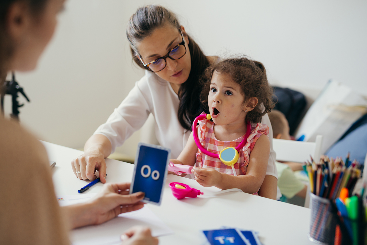 "A child practices pronouncing language sounds while sitting at a table with two adults"