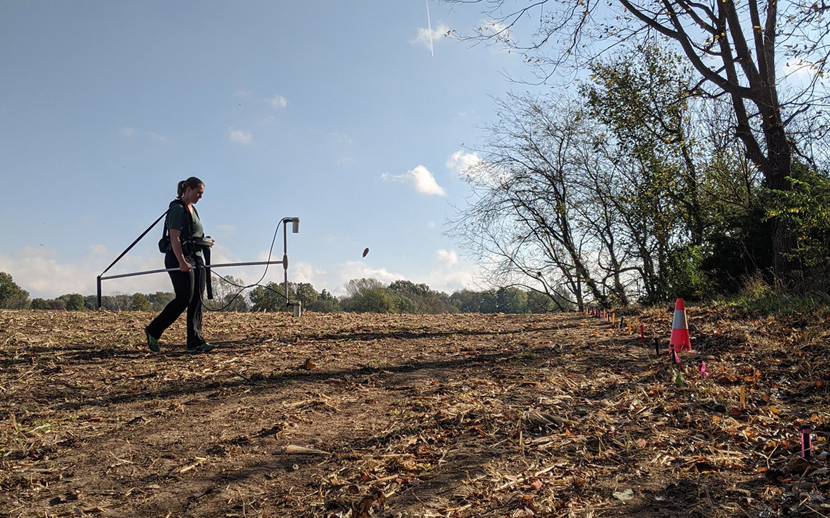 Blair Schneider carries scientific equipment through a field to collect data.