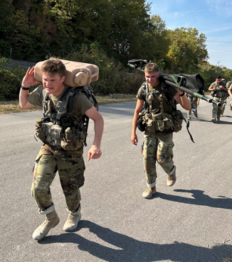 Cadets Jakob Lord, Oskaloosa, and Nate Lundgren, Olathe, carry heavy equipment on their shoulders on a sunny road.