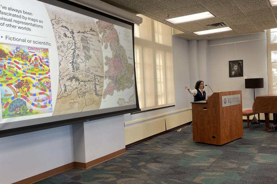 woman presents from podium, gesturing toward screen
