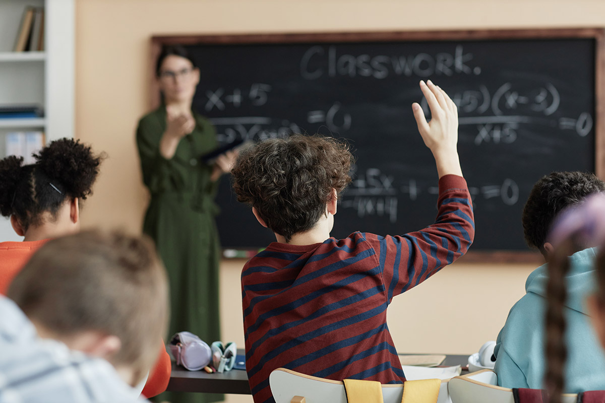 Seen from the back of a classroom, a student raises his hand as the teacher calls on him from the chalkboard.