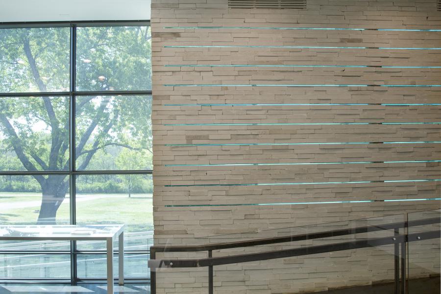 Interior view of the University of Kansas Center for Design Research showing a textured stone wall and large windows overlooking lush trees outside.