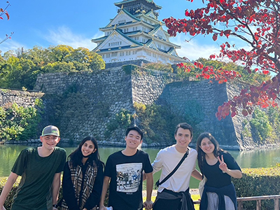 A group of five people stands smiling in front of Osaka Castle. The scene includes a clear blue sky, trees with autumn foliage, and a stone wall surrounding the castle.