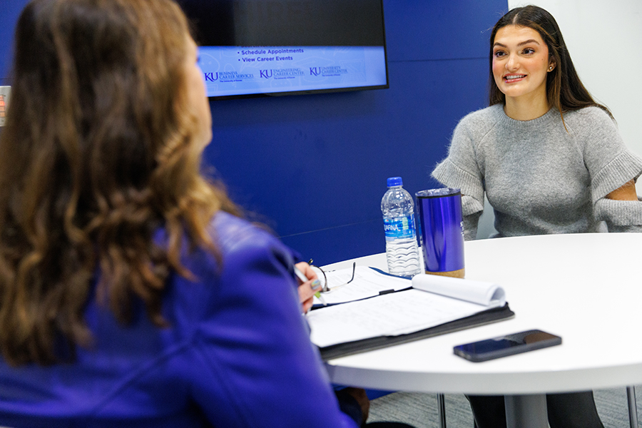 A woman with brown hair wearing a blue shirt sits facing a table with bottles of water and notebooks as girl in a gray sweater speaks.