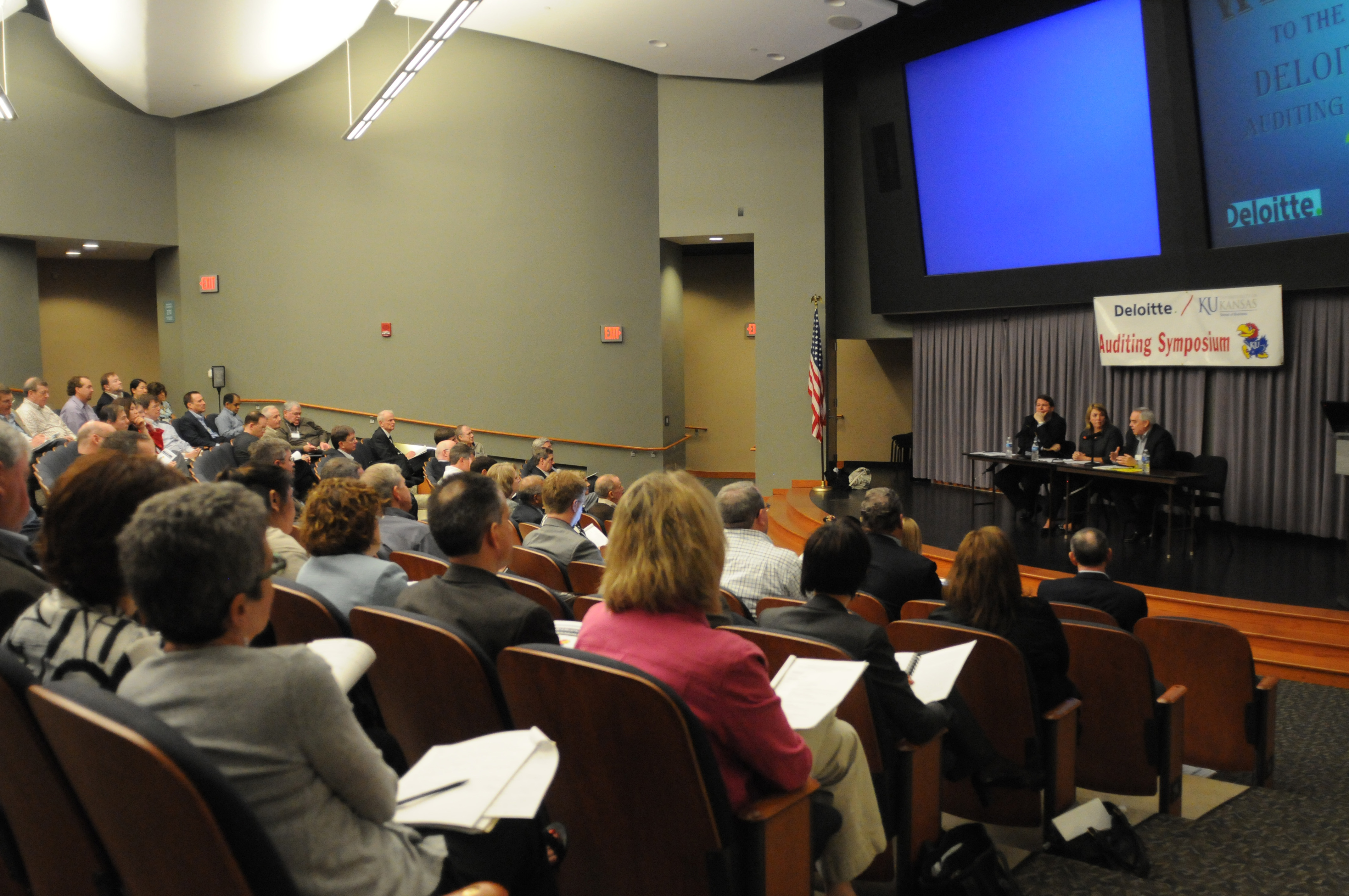 Audience members seated in a large auditorium during the Deloitte/KU Auditing Symposium, listening to panelists speaking at the front.