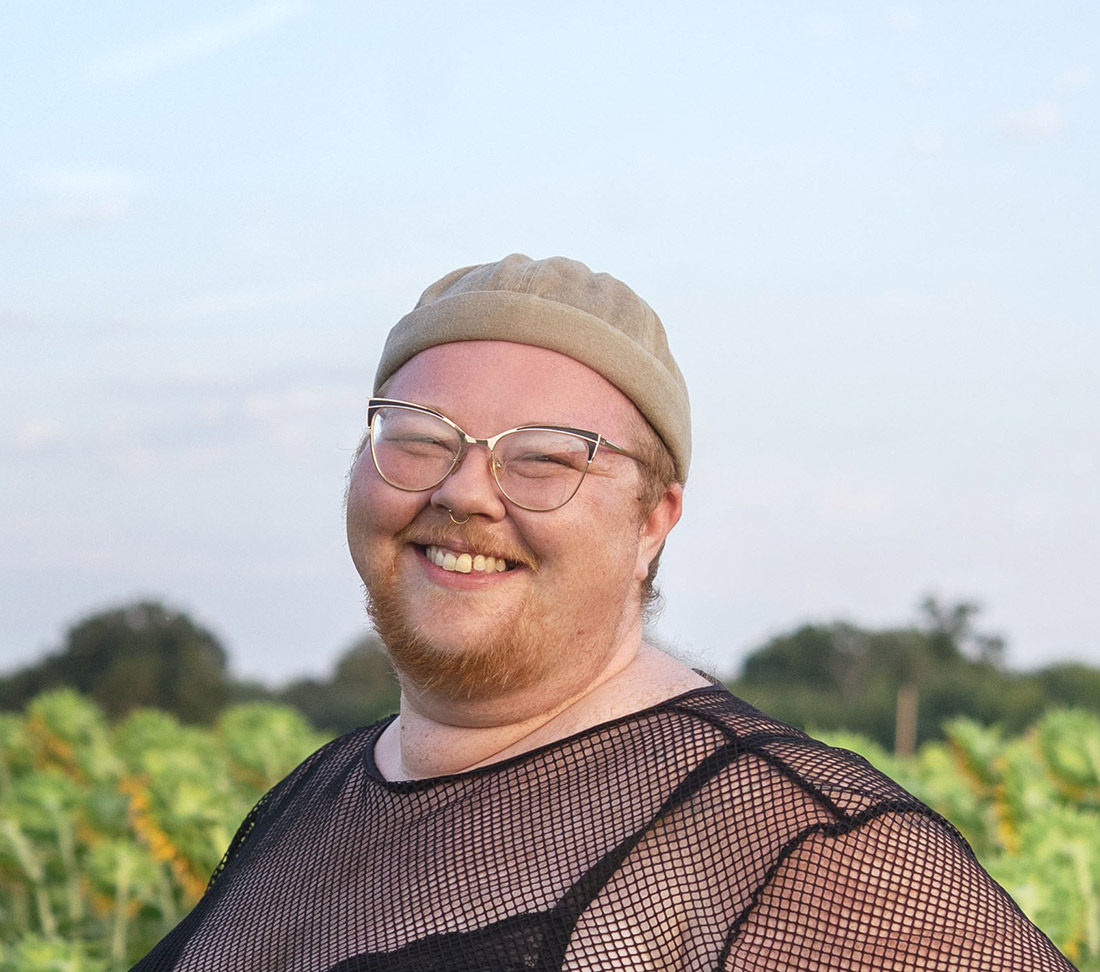 Arlowe Sue Clementine standing in a sunflower field and smiling widely. They wear metal rimmed glasses and a stocking hat