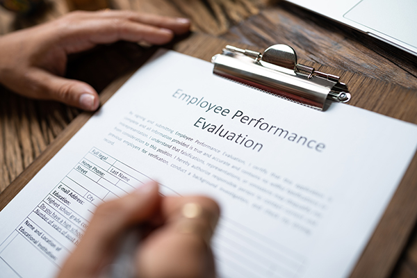 A person is writing on an "Employee Performance Evaluation" form placed on a clipboard. The form includes sections for ratings and comments. The clipboard rests on a wooden surface.