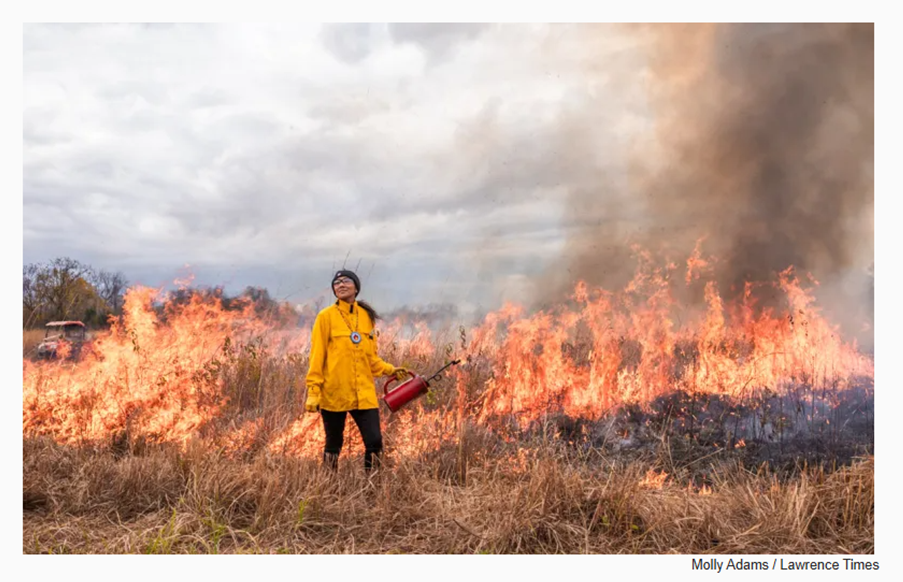 Melinda Adams wearing a yellow coat and holding a flame thrower stands in front of burning brush.